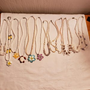 Peut inclure: Un ensemble de 12 colliers en coquillages blancs avec différents pendentifs, dont des fleurs, une tortue et une petite coquille. Les colliers sont présentés sur une surface blanche.