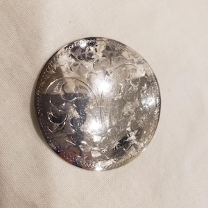 Peut inclure: Un objet rond en métal argenté avec un motif floral et de feuilles complexe. Le motif est gravé sur la surface, créant un aspect détaillé et texturé. L'objet a une qualité brillante et réfléchissante.