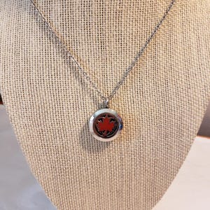 May include: A silver chain necklace with a round silver pendant featuring a red maple leaf design. The pendant is a diffuser for essential oils.