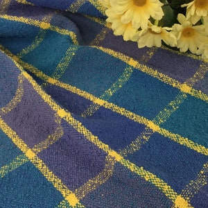 May include: A blue and purple plaid textile with yellow accent lines. The fabric is folded, with a textured appearance. Yellow flowers are placed in the upper right corner of the image.