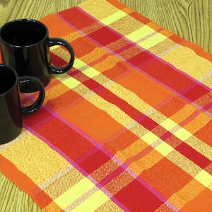 May include: Two black ceramic mugs with handles on a red, orange, and yellow plaid tablecloth.