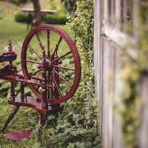 K&ouml;nnte beinhalten: Ein h&ouml;lzernes Spinnrad mit dunkelroter Oberfl&auml;che und einem gro&szlig;en, runden Rad. Das Rad steht auf einem dreif&uuml;&szlig;igen Holzgestell. Das Rad befindet sich in einem grasbewachsenen Bereich mit gr&uuml;nem Laub im Hintergrund.