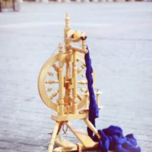 May include: A wooden spinning wheel with a blue yarn skein draped over it. The wheel is set against a backdrop of an arched building with a sign that reads "The Minstrel KROMSKI & SONS".