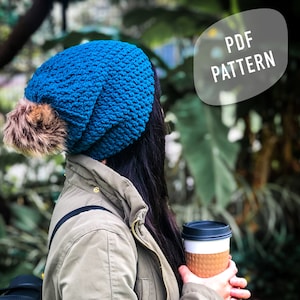 May include: A blue crocheted beanie with a fluffy pom pom. The beanie is worn by a person holding a takeaway coffee cup. The text "PDF PATTERN" is visible in the background.