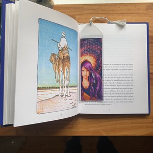May include: An open book with a colourful illustration of a camel and rider on the left page. A clear bookmark with a woman and geometric design is in the book. The bookmark has a grey tassel.
