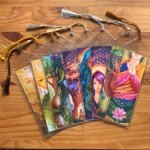 May include: A selection of illustrated bookmarks with vibrant artwork and decorative tassels. The bookmarks showcase diverse designs, including celestial and nature-inspired imagery. Each bookmark is encased in a clear plastic sleeve.