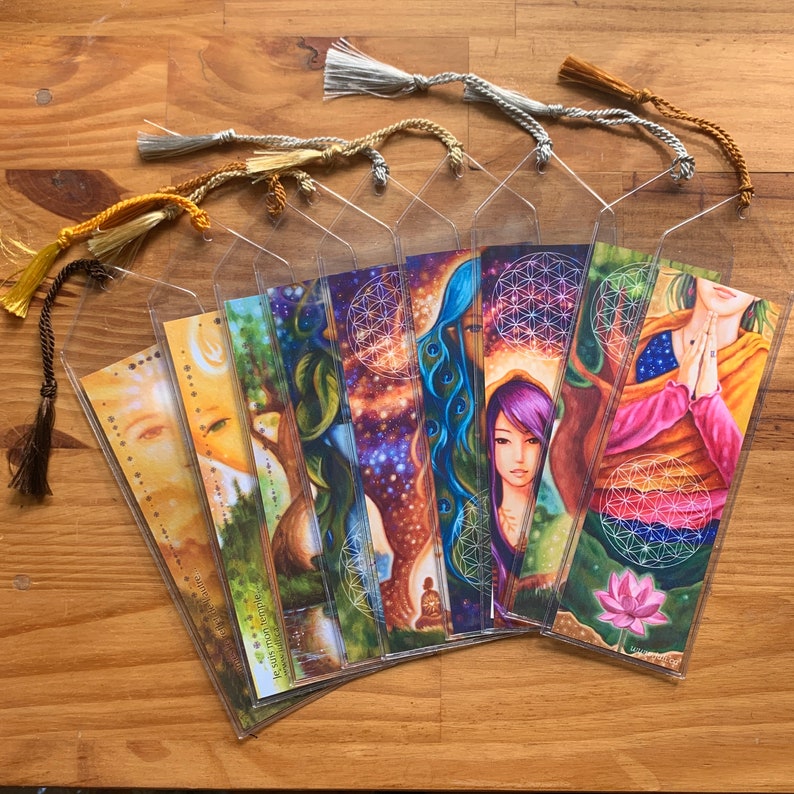 May include: A selection of illustrated bookmarks with vibrant artwork and decorative tassels. The bookmarks showcase diverse designs, including celestial and nature-inspired imagery. Each bookmark is encased in a clear plastic sleeve.