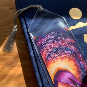 May include: A clear acrylic bookmark with a grey tassel rests on a navy blue book. The bookmark features a colourful image of a galaxy with a white geometric design. A unique accessory for book lovers.