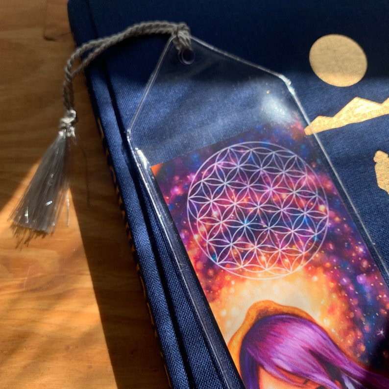 May include: A clear acrylic bookmark with a grey tassel rests on a navy blue book. The bookmark features a colourful image of a galaxy with a white geometric design. A unique accessory for book lovers.