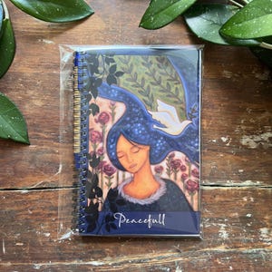 May include: A spiral-bound notebook featuring a colourful illustration of a person with long blue hair, a white dove, and pink roses. The cover displays the word "Peacefull" in white script at the bottom, all encased in a clear plastic protective sleeve.