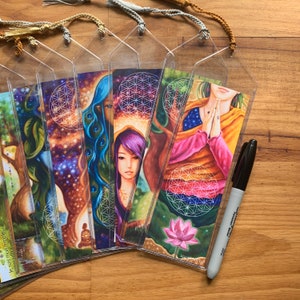 May include: A selection of vibrant bookmarks with spiritual artwork, featuring women, trees, and cosmic designs. Each bookmark has a clear plastic sleeve and a decorative tassel. A black marker is also present.