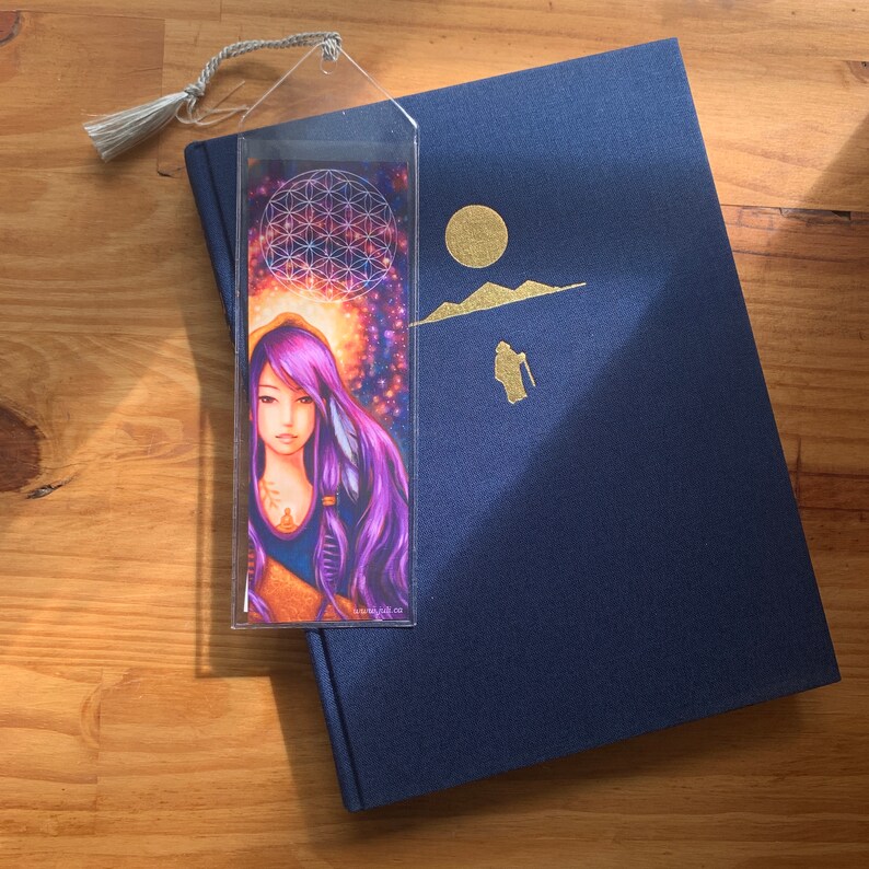 May include: A navy blue journal with gold foil accents of a sun, mountains, and a person walking. A clear bookmark with a woman with purple hair and a geometric design hangs from the top of the journal. The bookmark has a tassel.
