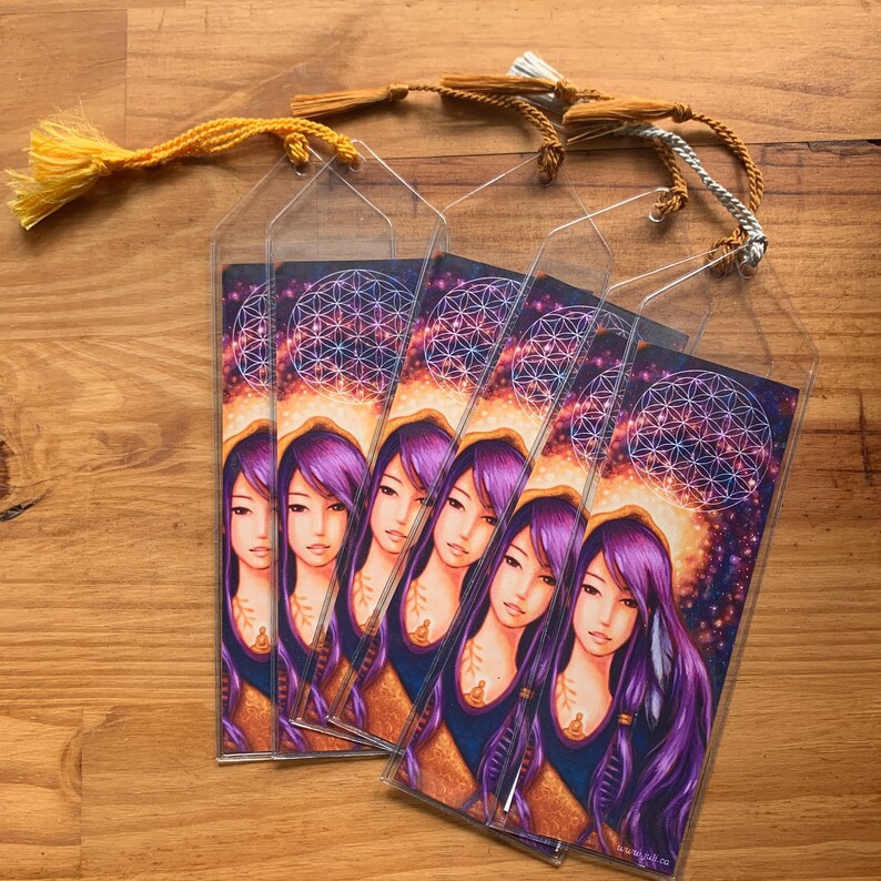 May include: Several clear bookmarks with a portrait of a woman with purple hair and a cosmic background. Each bookmark has a tassel attached. The woman's portrait is set against a backdrop of a geometric design.