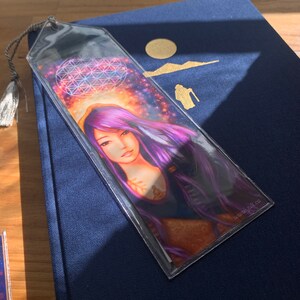 May include: A clear bookmark with a woman with purple hair and a celestial background. The bookmark has a tassel and the text "www.judi.ca". The artwork includes a geometric design and a sun symbol.