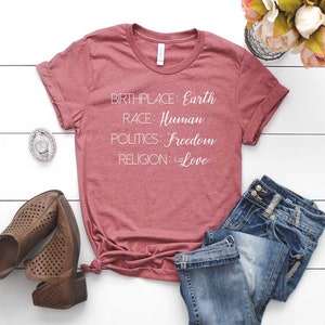 May include: A maroon t-shirt with the words "BIRTHPLACE: Earth, RACE: Human, POLITICS: Freedom, RELIGION: Love" in white script. The shirt is paired with blue jeans and brown cutout heeled shoes.