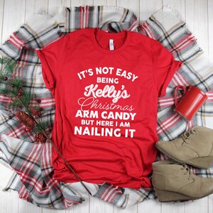 May include: A red t-shirt with the text "IT'S NOT EASY BEING Kelly's Christmas ARM CANDY BUT HERE I AM NAILING IT" in white script. The shirt is laid on a plaid blanket with a sprig of pine and a red mug and tan wedge boots.