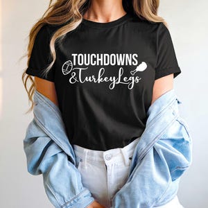 May include: Black t-shirt featuring the white text "TOUCHDOWNS & Turkey Legs". The design includes a football and a turkey leg graphic. The shirt is a casual style.