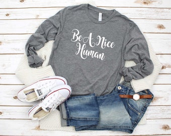 May include: Gray long-sleeve t-shirt with the text "Be A Nice Human" in white lettering. The shirt is paired with blue jeans, white sneakers, and a white cardigan.
