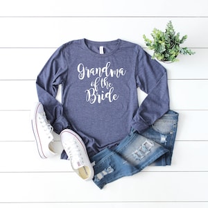 May include: A navy blue long-sleeved shirt with the text "Grandma of the Bride" in white lettering. The shirt is laid flat on a white wooden surface with a pair of white sneakers and blue jeans.