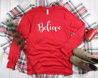May include: A red long-sleeve shirt with the word "Believe" in white script. The shirt is laid on a plaid blanket with a sprig of greenery, a red mug, and a pair of tan suede shoes. The items are arranged on a white wooden surface.