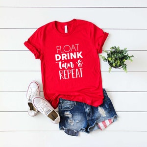 May include: A red t-shirt with the words "FLOAT DRINK Tan & REPEAT" in white lettering, paired with denim shorts, white sneakers, and a small potted plant. The items are arranged on a white wooden surface.
