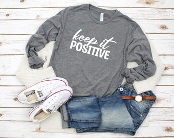 May include: A heather grey long-sleeved t-shirt with the text "keep it positive" in white lettering. The shirt is styled with white sneakers, blue jeans, and a white cardigan.