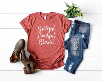 Puede incluir: Una camiseta de color óxido con las palabras "Grateful, Thankful, Blessed" en escritura blanca. La camiseta se combina con vaqueros azules desgastados, botines marrones y una pequeña planta en maceta sobre una superficie de madera blanca.