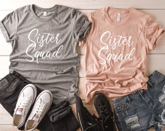 May include: Two gray and peach t-shirts with the text "Sister Squad" in white cursive lettering. The shirts are laid out on a white wooden surface with jeans and sneakers.