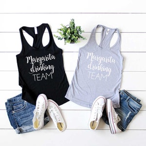 Puede incluir: Dos camisetas sin mangas, una negra y otra gris, con el texto "Margarita drinking TEAM" en escritura blanca. Se muestran con pantalones cortos vaqueros y zapatillas blancas. También se ve una pequeña planta en maceta.
