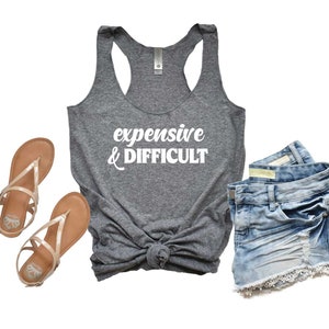 Könnte beinhalten: Ein graues Tanktop mit dem Text "expensive & difficult" in Weiß. Das Tanktop wird mit Jeans-Shorts und Sandalen kombiniert.