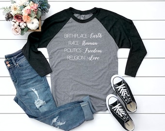 May include: A gray and black raglan long-sleeve shirt with the text "BIRTHPLACE: Earth, RACE: Human, POLITICS: Freedom, RELIGION: Love." The image also includes blue jeans, black sneakers, and a floral arrangement.