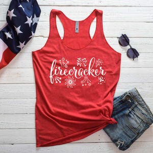 May include: A red racerback tank top with the word "firecracker" in white script and fireworks graphics. The tank top is paired with blue denim shorts and sunglasses, with an American flag in the background.