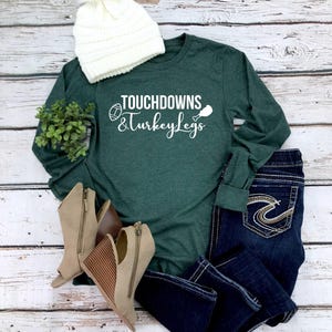 May include: A casual outfit comprising a dark green long-sleeved top with the text "Touchdowns & Turkey Legs", dark wash jeans, beige wedge booties, and a white knitted beanie with a pom-pom. A small potted plant is also included.