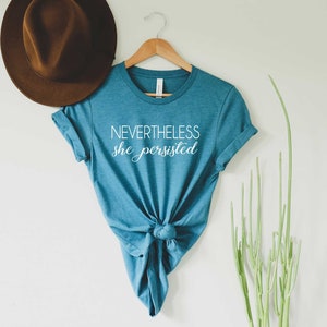 May include: A teal blue t-shirt with the text "NEVERTHELESS she persisted" in white lettering. The shirt is tied in a knot at the bottom.