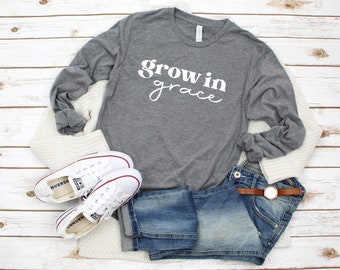 May include: Gray long-sleeve t-shirt with the text "grow in grace" in white lettering. The shirt is styled with white sneakers, blue jeans, and a white cardigan.