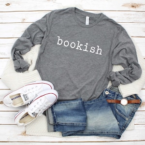 May include: A gray long-sleeve shirt with the word "bookish" in white, paired with blue denim shorts, white sneakers, and a cream-colored sweater. A brown leather belt and a watch complete the outfit.