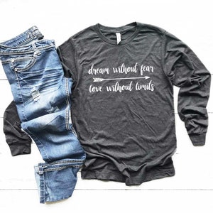 May include: A dark gray long-sleeve shirt and blue jeans are displayed on a white wooden surface. The shirt has the words "dream without fear, love without limits" printed in white. The jeans are blue denim.