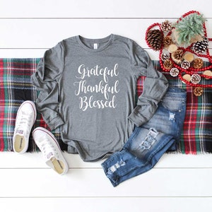 K&ouml;nnte beinhalten: Ein graues Langarmshirt mit dem Schriftzug "Grateful Thankful Blessed" in wei&szlig;er Schrift. Das Shirt wird mit zerrissenen blauen Jeans und wei&szlig;en Turnschuhen kombiniert. Eine karierte Decke und dekorative Tannenzapfen vervollst&auml;ndigen das Ensemble.