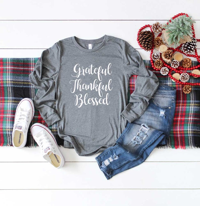 K&ouml;nnte beinhalten: Ein graues Langarmshirt mit dem Schriftzug "Grateful Thankful Blessed" in wei&szlig;er Schrift. Das Shirt wird mit zerrissenen blauen Jeans und wei&szlig;en Turnschuhen kombiniert. Eine karierte Decke und dekorative Tannenzapfen vervollst&auml;ndigen das Ensemble.