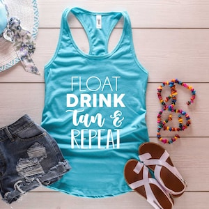May include: A turquoise tank top with the words "FLOAT DRINK Tan & REPEAT" in white script. The image also includes denim shorts, a sun hat, a beaded necklace, and sandals, all arranged on a light wood-grain surface.