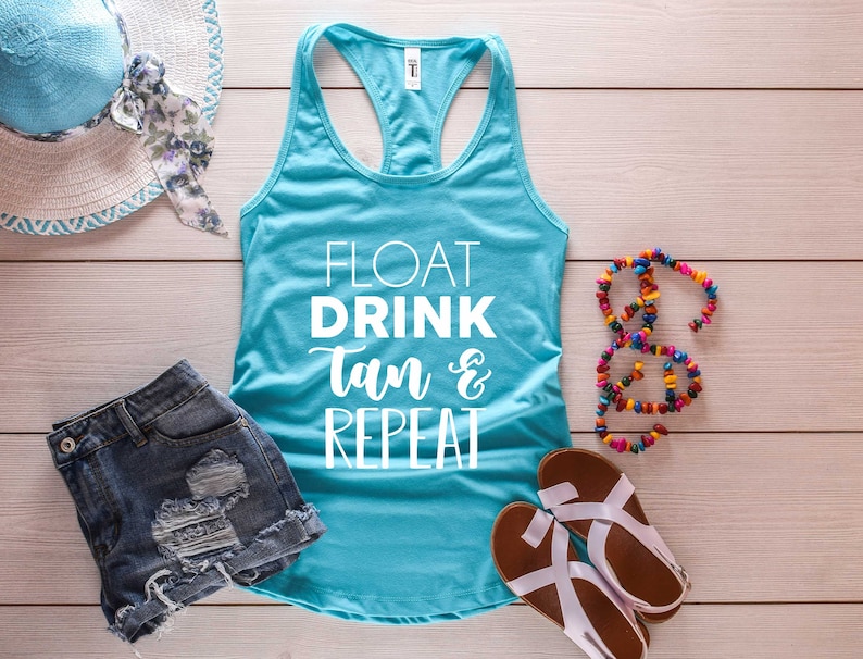 May include: A turquoise tank top with the words "FLOAT DRINK Tan & REPEAT" in white script. The image also includes denim shorts, a sun hat, a beaded necklace, and sandals, all arranged on a light wood-grain surface.