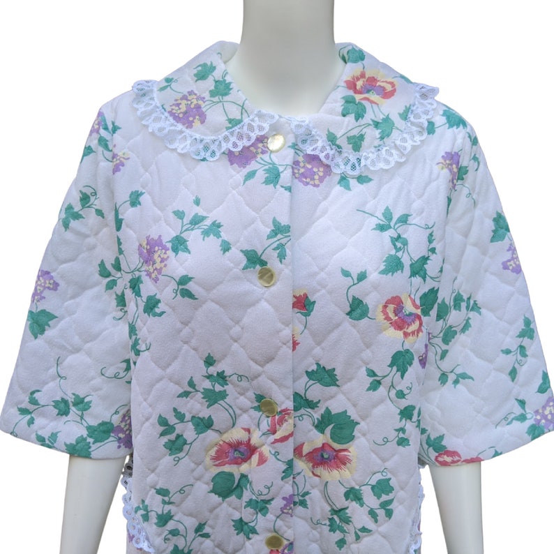 Puede incluir: Una bata blanca acolchada con estampado floral y cuello de encaje. La bata tiene cierre de botones en la parte delantera y mangas cortas.