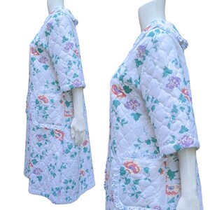 Puede incluir: Una bata acolchada blanca con estampado floral con flores rosas, moradas y verdes. La bata tiene un ribete de encaje alrededor de los bordes y bolsillos.