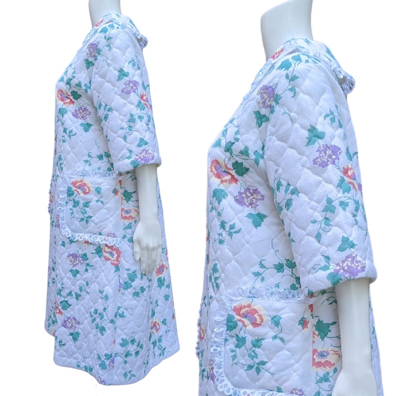 Puede incluir: Una bata acolchada blanca con estampado floral con flores rosas, moradas y verdes. La bata tiene un ribete de encaje alrededor de los bordes y bolsillos.