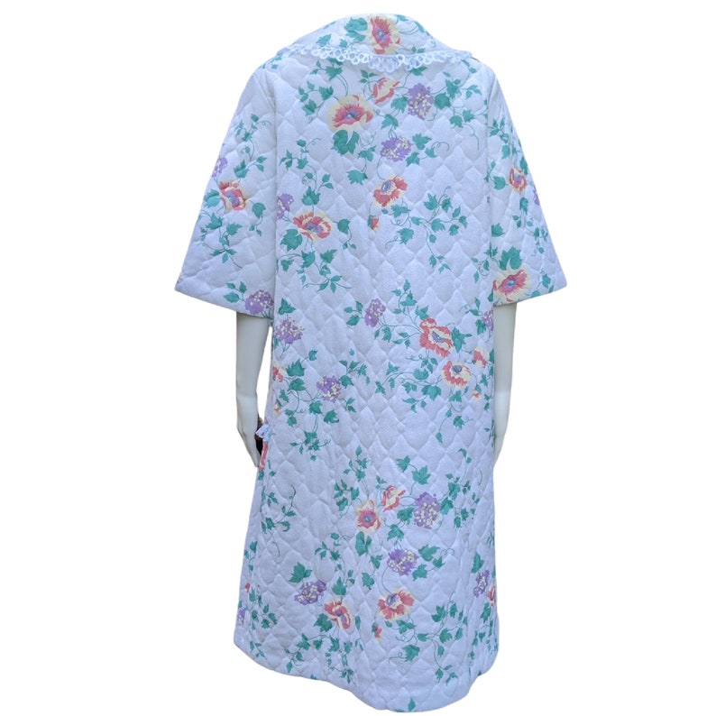 Puede incluir: Una bata acolchada blanca con estampado floral con flores rosas, naranjas y moradas. La bata tiene un cuello con ribete de encaje y mangas cortas.