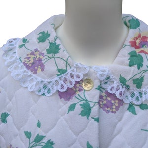 Puede incluir: Batista blanca acolchada con estampado floral con hojas verdes, flores moradas y detalles amarillos. La bata tiene un ribete de encaje blanco alrededor del cuello y un bot&oacute;n dorado.