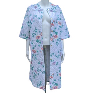 Puede incluir: Una bata acolchada blanca con estampado floral y ribete de encaje. La bata tiene cierre de botones y mangas cortas.