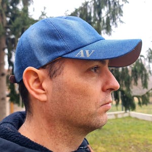 Può includere: Un cappello da baseball in denim blu con visiera marrone. Il cappello ha le iniziali "AV" ricamate sul davanti in colore argento.