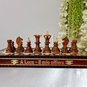 Puede incluir: Un juego de ajedrez de madera con un tablero de ajedrez a cuadros marrón y blanco, las piezas están dispuestas en la posición inicial. El tablero de ajedrez está en una caja de madera con un interior rojo y la inscripción "A Luca - Il mio alfiere" en la parte delantera.