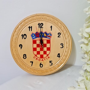 Puede incluir: Un reloj de madera con una esfera blanca y números negros. El reloj tiene un escudo de armas croata en el centro con la palabra "CROATIA" impresa debajo.